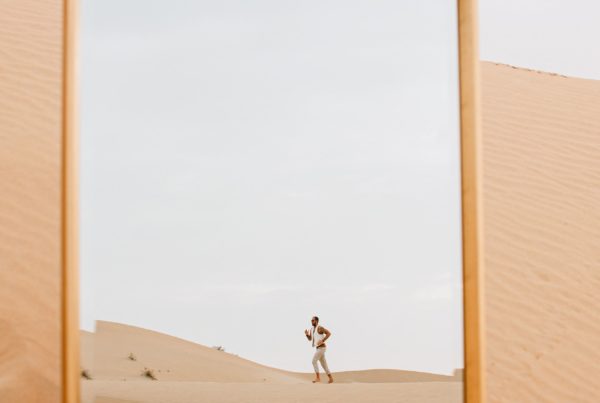 A person stands at a distance in the desert, framed by a large mirror positioned in the sand.