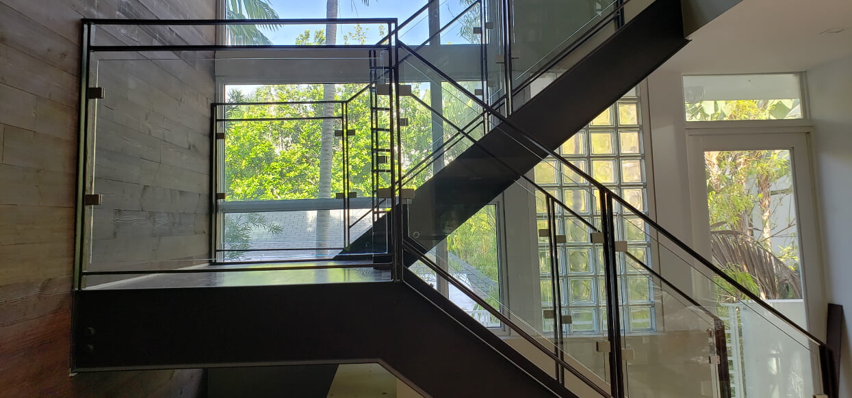 Modern staircase with custom glass panels and black metal railings in a bright room with large windows and wooden walls.