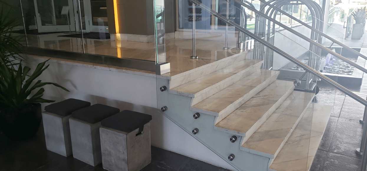 Modern lobby with custom glass marble stairs and a curved glass railing, flanked by a reception desk and seating stools.