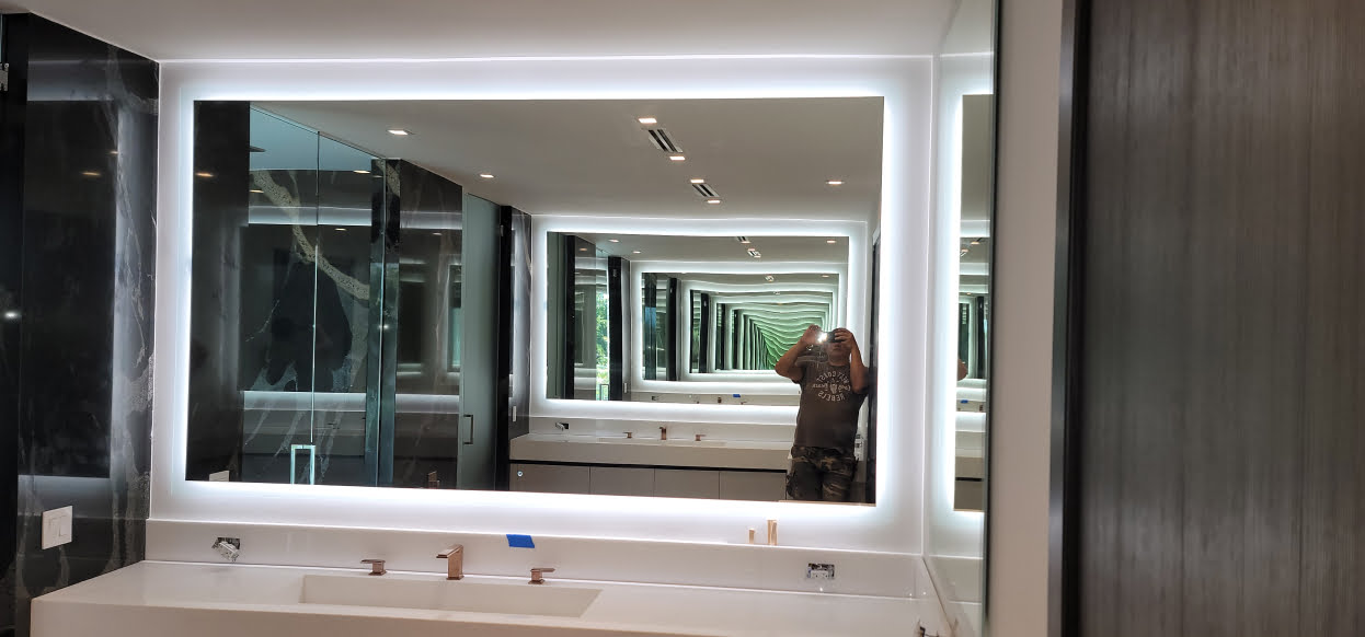 A modern bathroom featuring custom glass mirrors in Hollywood, with a large lit mirror casting a reflection of two sinks and numerous smaller mirrors creating an infinite reflection effect. A person is visible taking the photo.
