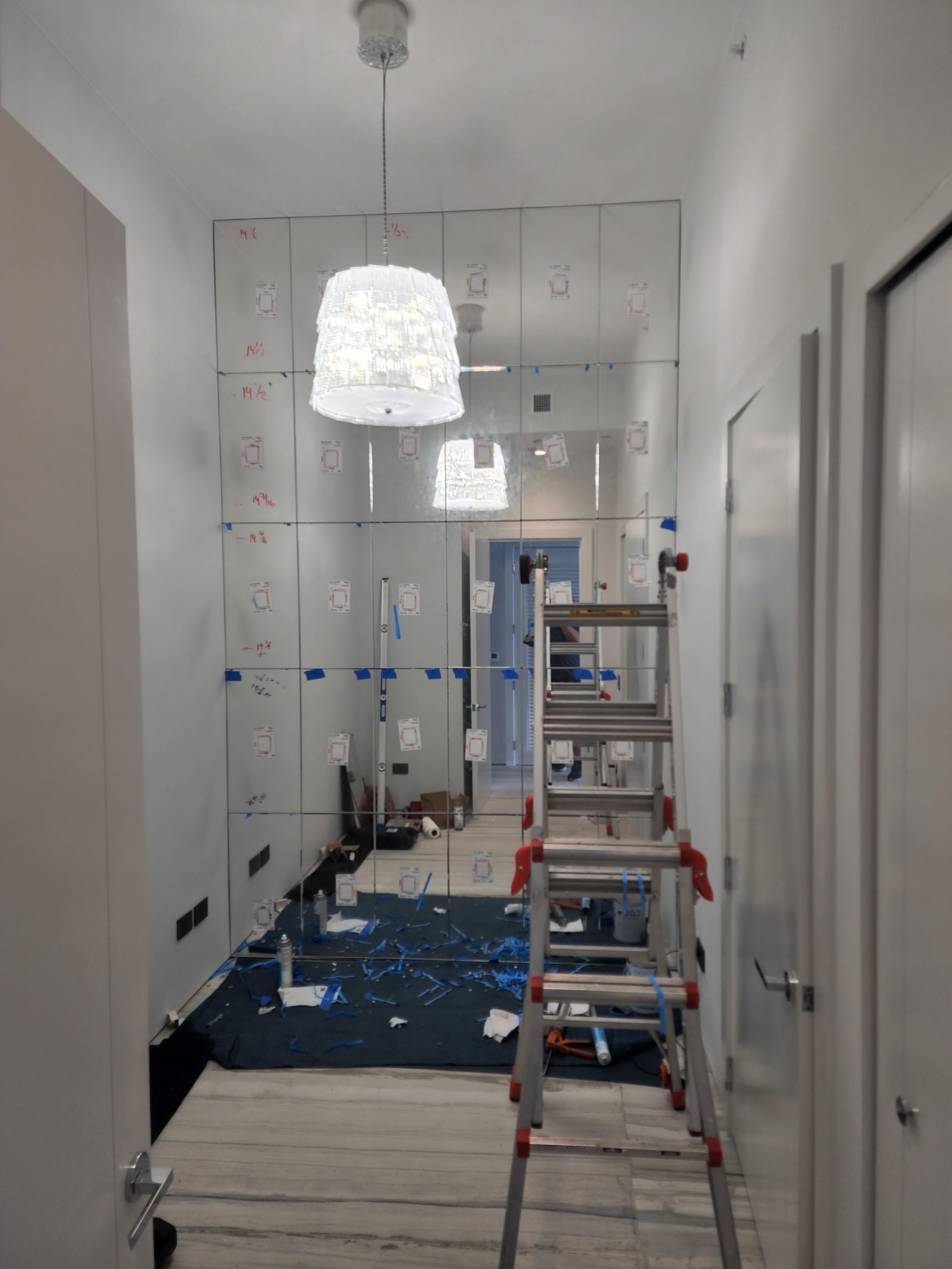 Interior of a room under renovation with custom glass & mirrors in Boca Raton, protective sheets on the floor, a ladder in the center, and walls marked for measurements.