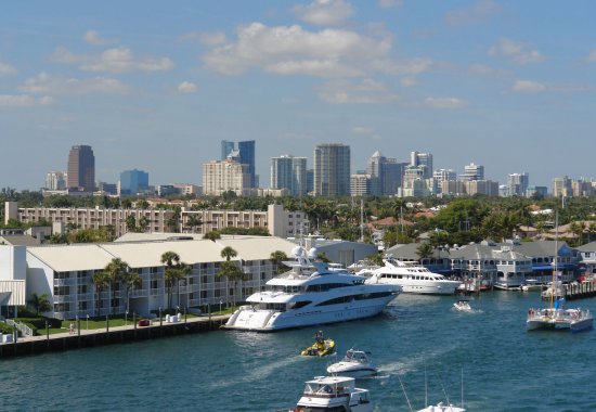 A view of Fort Lauderdale, Florida, featuring custom glass and mirrors on luxury yachts along the waterway, with a modern cityscape and clear blue sky in the background.