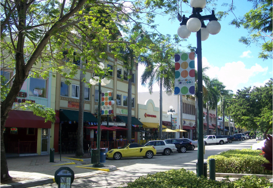 A sunny street scene showing a row of colorful, multi-story buildings with various shops, including a custom glass & mirrors shop in Boca Raton, a yellow car parked, and a lamppost with