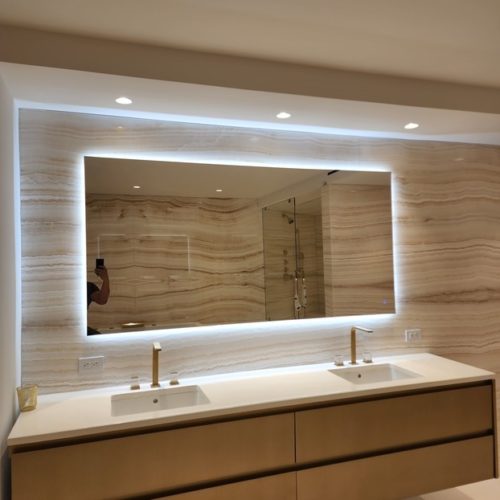 Backlit mirror in a modern bathroom with double sink and marble wall. Built-in lights and gold faucets enhance the elegant design.