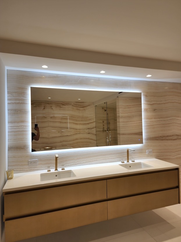 Backlit mirror in a modern bathroom with double sink and marble wall. Built-in lights and gold faucets enhance the elegant design.