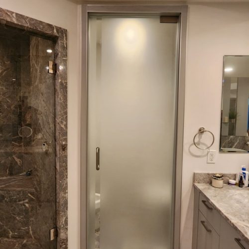 Modern bathroom with frosted glass partitions and marble finishes. The glass door leads to a shower area with marble detailing, next to a sink and mirror.